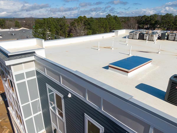 Rooftop view of a flat white roof with a blue skylight, windows along the edge, and HVAC units in the distance under a blue sky.